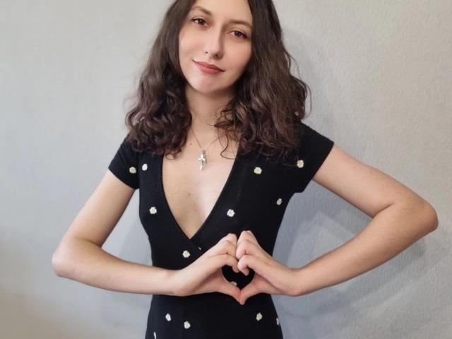Nichole Kanzenbach, mit welligem, braunem Haar, kleinen Brüsten, trägt ein schwarzes Kleid mit Punkten, formt mit ihren Händen ein Herz, lächelt und trägt eine Halskette..