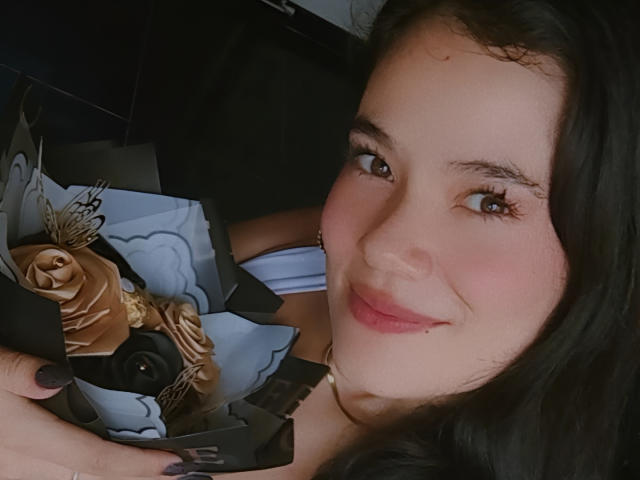 IsabellaVoss with dark hair, brown eyes, and pink lips, holding a bouquet of roses and chocolate, smiling softly at the camera. Her small, cute nose is slightly upturned.