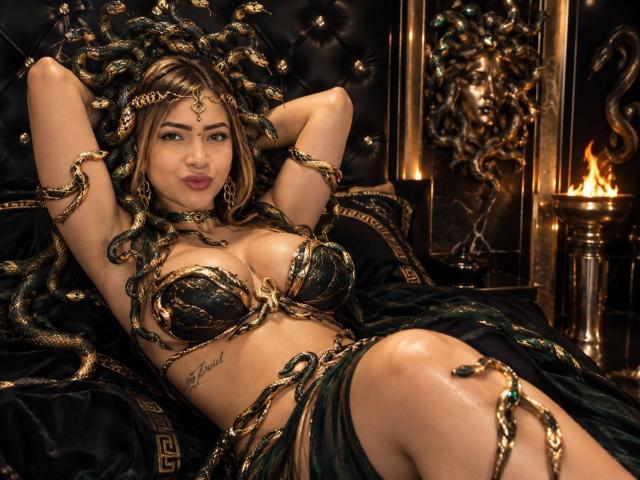 Tatiana Rossi mit grün-goldenen, schlangenartigen Schmuckstücken, großen Brüsten, Tattoo auf der rechten Seite, liegend auf schwarzem Samt, Arme hinter dem Kopf, grüne und schwarze Dessous, Feuer im Hintergrund..