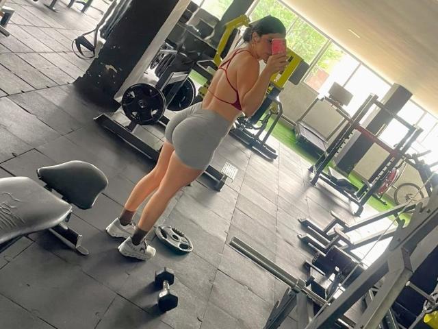 VictoriaFiore with a red sports bra and gray booty shorts, standing in a gym, taking a mirror selfie, showing off her ass and white sneakers.