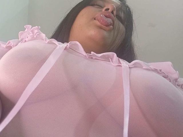 KonyBella com a língua de fora, vestindo uma blusa rosa transparente, mostrando seios grandes e mamilos visíveis, fotografada de um ângulo baixo. Seu cabelo escuro e pele clara são visíveis..