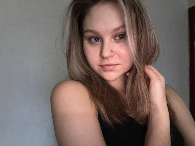 YungQuiller with light brown, slightly messy hair, small nose ring, blue eyes, fair skin, and a subtle smile, wearing a black top, hand near face.