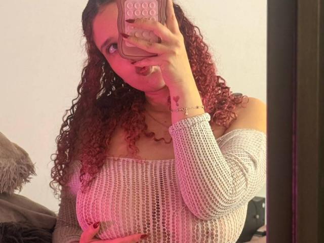 ValeryScot con capelli rossastri ricci, che tiene un telefono, indossa una maglia bianca a maglia con una spalla scoperta, mostrando il suo seno prosperoso, luce rosa, selfie allo specchio, custodia rosa per il telefono, anello alla mano destra..