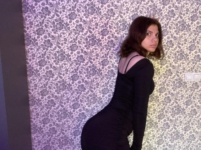 CicelyDence with wavy brown hair, black off-shoulder dress, and curvy ass, standing with back to camera, looking over shoulder. Patterned wallpaper behind her.