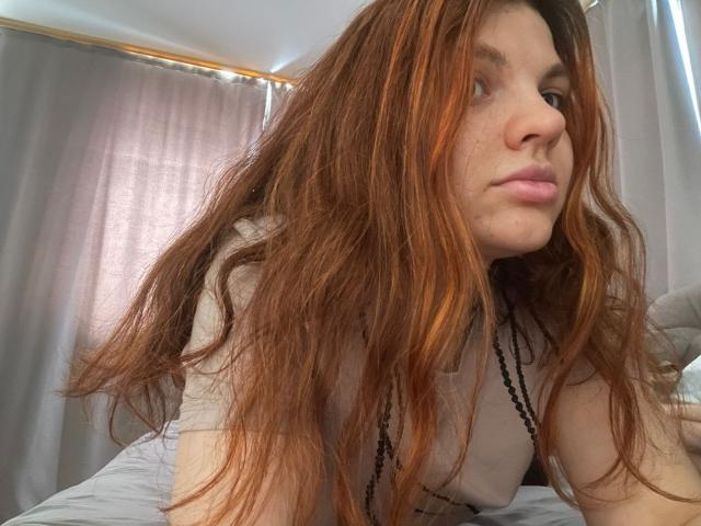 Cicely Dence hat langes, zerzaustes rotes Haar, eine niedliche Nase und rosafarbene Lippen. Sie trägt ein beiges Hemd mit einer schwarzen Halskette und blickt leicht zur Seite, mit einem leichten Schmollmund..