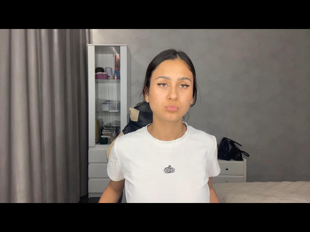 JamesPaino with straight brown hair, wearing a white tee, small black logo, neutral expression, standing in a room with gray curtains and white dresser.