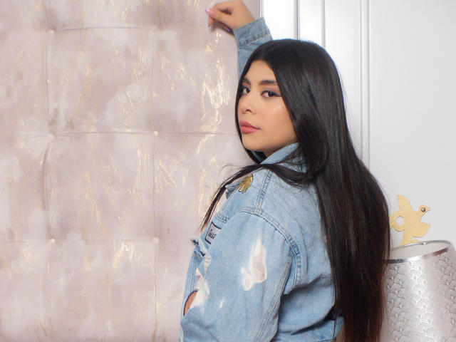 ArianaEvan with long black hair, wearing a blue denim jacket, looking over her shoulder, standing next to a silver lamp with a butterfly ornament.