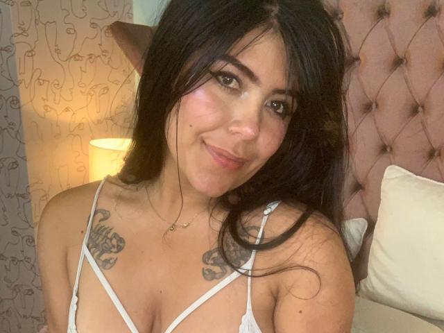 CatalinaReiyna with long black hair, brown eyes, and a slight smile, wearing a white strappy top, showing small tattoos on her tits, in a cozy bedroom.