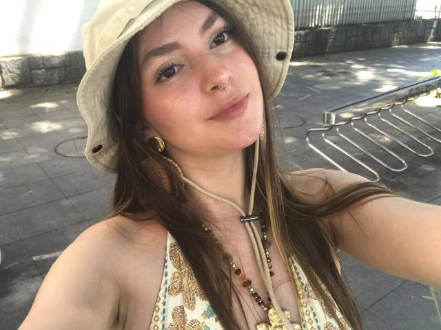 VelvetMoonx with long brown hair, wearing a beige bucket hat, nose ring, and gold earrings, showing her tits in a patterned top, smiling in a sunlit outdoor setting.