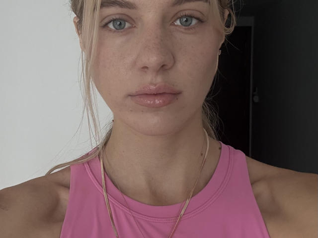 Lakeesha Janke tem lábios rosados brilhantes, olhos azuis e está vestindo uma blusa esportiva rosa com um colar dourado. Ela tem sardas claras e uma expressão neutra, com os lábios levemente entreabertos..