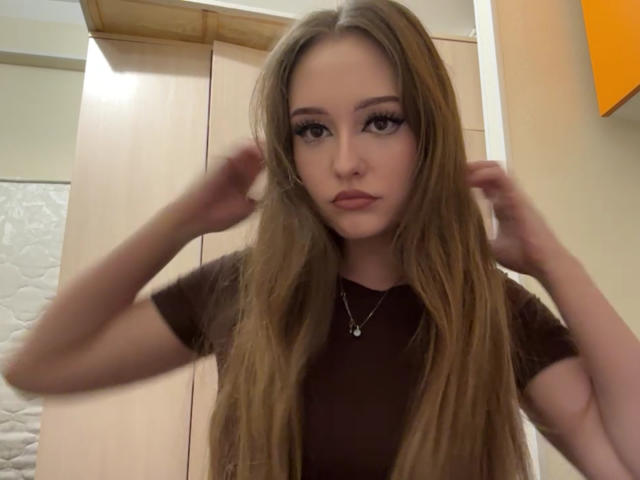 MinhMajic con lunghi capelli castani, indossa una camicia marrone, le mani tra i capelli, espressione seria, una piccola collana, pelle chiara, sopracciglia folte e un trucco leggero..