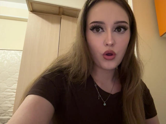 MinhMajic with long brown hair, big eyes, and red lipstick, wearing a black shirt, looking at the camera with a slightly open mouth, necklace with a small pendant.