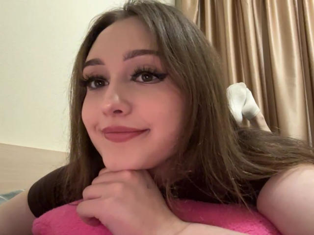 MinhMajic, met lang bruin haar, roze lippenstift en dikke wimpers, lachend, met haar kin op haar hand rustend, draagt een roze top, heeft bruine ogen en een schattig gezicht..
