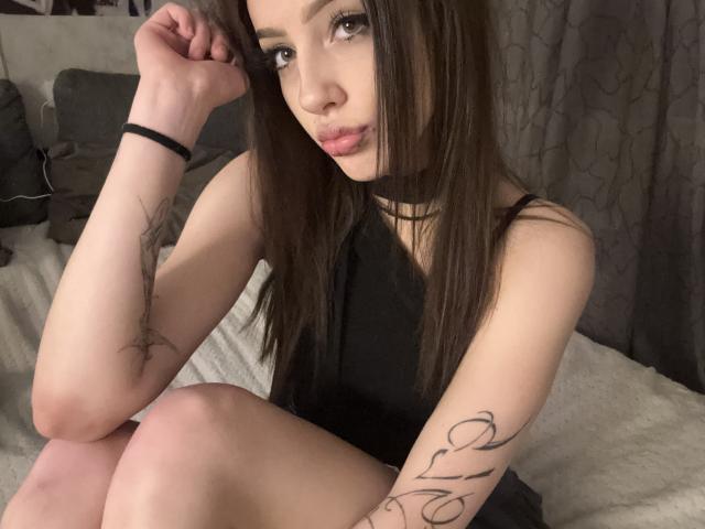 SimonneBrustkern with long brown hair, black choker, and black dress, showing tattoos on arms, sitting on bed, pouty lips, gray curtain background, and black wristband.