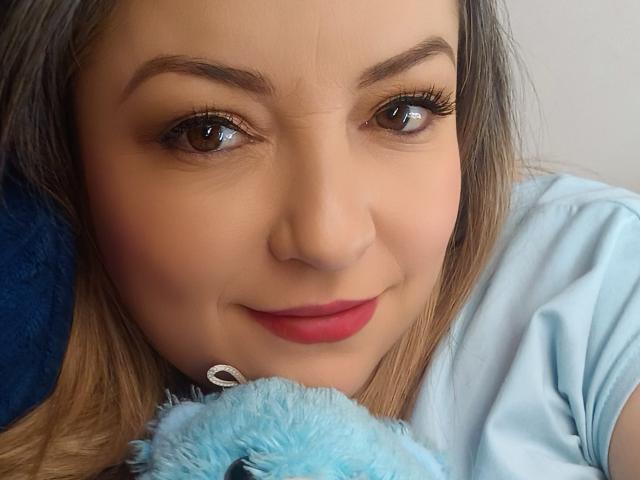 NatalyHami, con occhi marrone chiaro, rossetto rosso e un trucco leggero, tiene un morbido giocattolo blu, indossa una camicia azzurra e sorride dolcemente alla fotocamera..