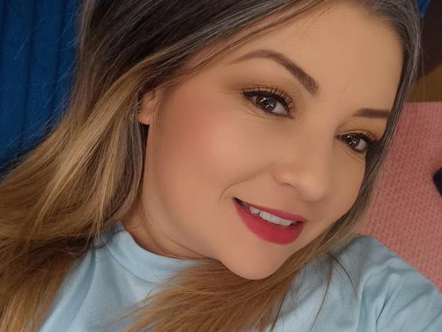 NatalyHami with blonde hair, light skin, red lipstick, and brown eyes, smiling at the camera, wearing a light blue shirt, and small ear piercing, in a selfie.