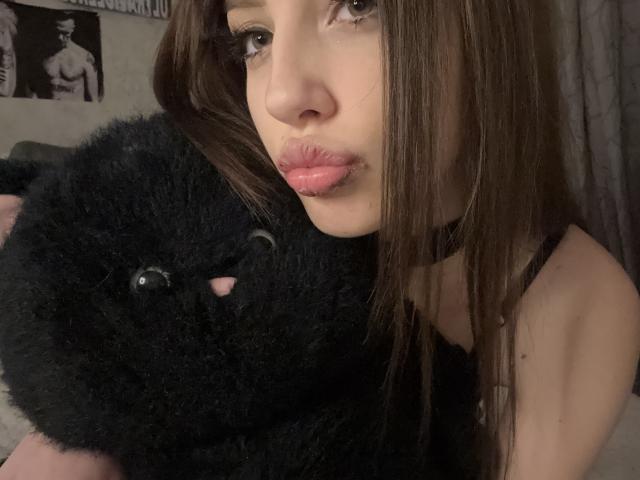 SimonneBrustkern with pouty pink lips, brown hair, holding a fluffy black stuffed cat, wearing a black choker, in a dimly lit room. Her eyes are half-open.