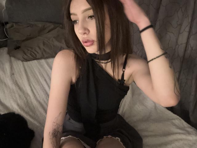 SimonneBrustkern with long brown hair, black dress, choker, and tattoo on right arm, sitting on bed, hand in hair, looking at camera, slightly open mouth, pale skin, black bra.