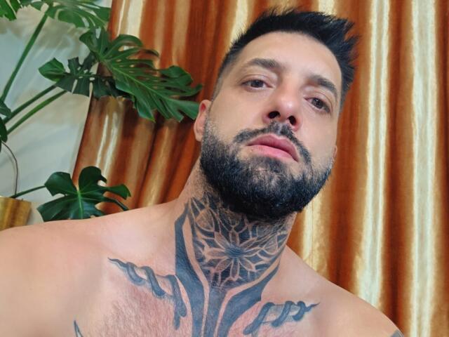 TommyDum, avec une barbe, des cheveux foncés et des tatouages sur le cou et la poitrine, regardant vers le bas la caméra. Une plante verte sur la gauche, des rideaux dorés en arrière-plan..