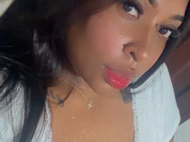 NatyLm's face with dark brown skin, shiny pink lips, black nose ring, small tattoo on neck, and gold cross necklace, wearing a light blue shirt, looking to the side.