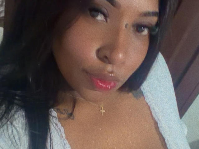 NatyLm with dark brown skin, long black hair, and red lipstick, wearing a blue top, a gold cross necklace, and a small beauty mark above her right eyebrow.