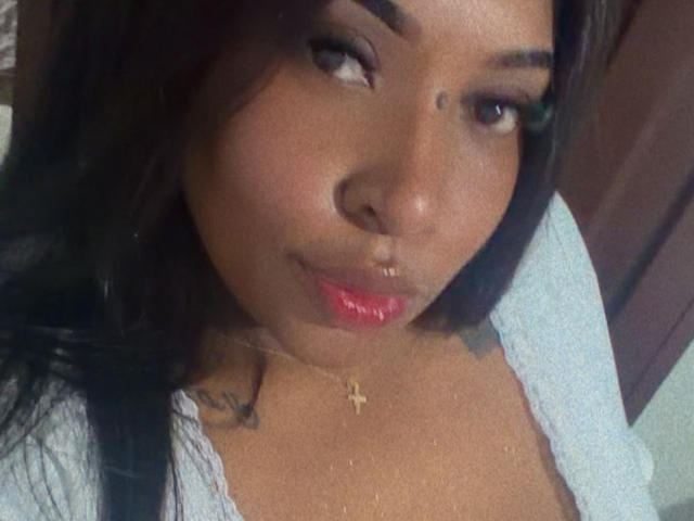 NatyLm's face, brown skin, long black hair, small nose, red lipstick, slight smile, gold necklace with cross, visible tattoo on left shoulder, wearing light blue top.