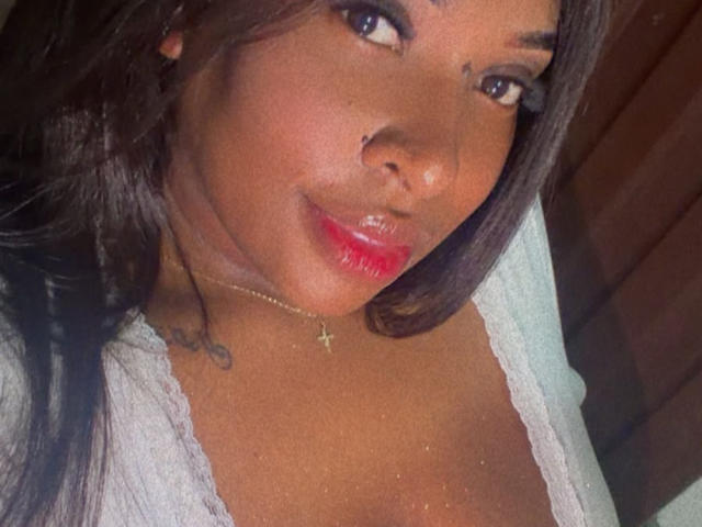 NatyLm with shiny red lips, dark brown skin, and long black hair, wearing a white lace top, has a cross necklace and a small tattoo on her left shoulder.