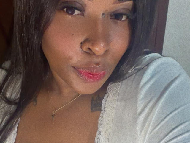 NatyLm with dark brown skin, long black hair, nose ring, and red lipstick, wearing a white lace-trimmed top, showing small tattoos on her shoulders.