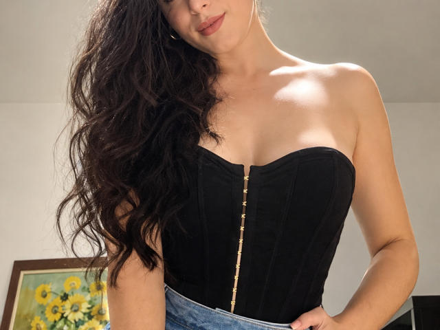 VickyMegan with wavy dark hair, wearing a black strapless corset with a gold zipper, showing her tits, in a selfie-style photo. She's got a subtle smile.
