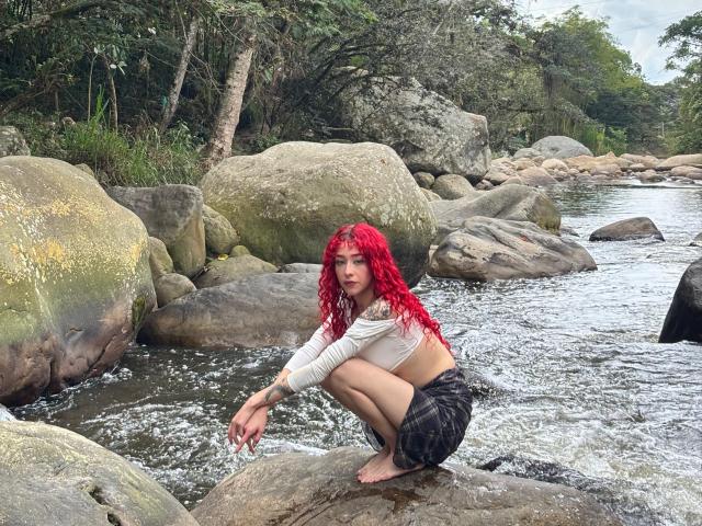 GaiaDoll with red hair squatting on a rock in a river, wearing a white long-sleeve top and black plaid skirt, her bare feet in the water, looking at camera.