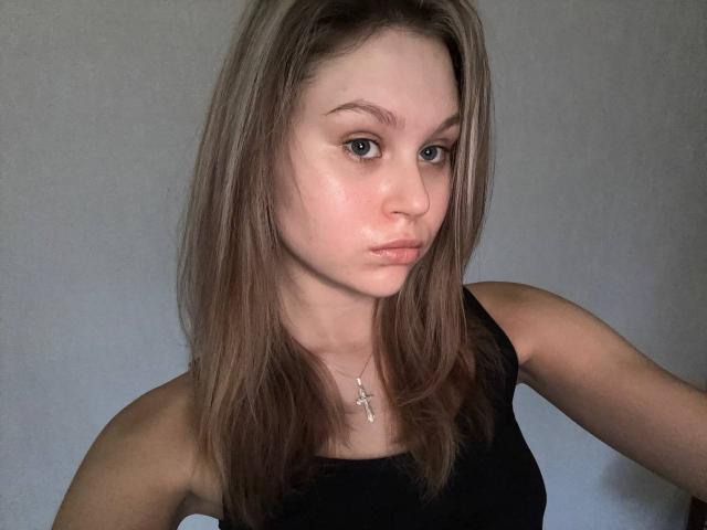 YungQuiller has light brown hair, blue eyes, and a cross necklace. She's wearing a black sleeveless top, looking serious with a slight pout. Her tits are small.
