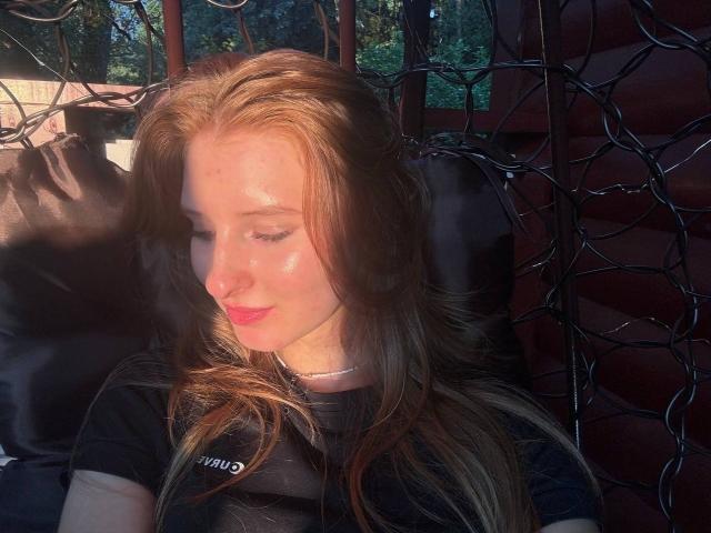 ArdelleCooksey ha una pelle luminosa, baciata dal sole, con capelli rossi, occhi chiusi e labbra rosa, indossa una maglietta nera ed è seduta contro dei fili scuri e aggrovigliati. Il suo viso è illuminato dalla luce del sole..