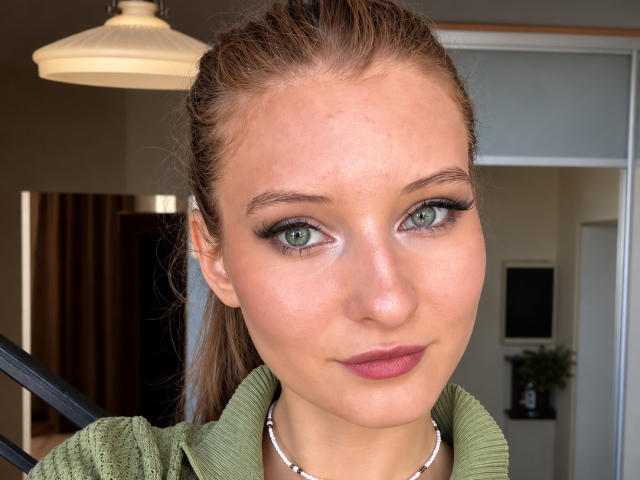ArdelleCooksey's face, showing green eyes, dark eyeliner, pink lipstick, and brown hair in a ponytail, wearing a green top and silver necklace. She's smiling slightly.