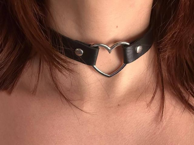 Le cou de SpencerLe est orné d'un collier en cuir noir avec un anneau métallique en forme de cœur, laissant apparaître quelques mèches de cheveux bruns et ondulés, ainsi qu'une peau claire..