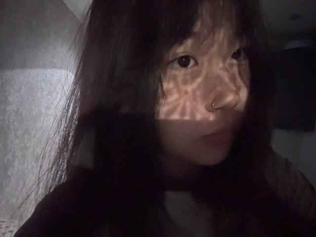 JungMcgranor with dark, messy hair and a nose ring, showing shadowy, geometric patterns on her face from light. She has a slight smile, looking to the side.