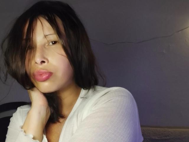 Lalala69, avec ses cheveux foncés et en désordre, ses lèvres roses et pulpeuses, et une chemise blanche texturée, repose sa tête sur sa main dans une pièce faiblement éclairée..