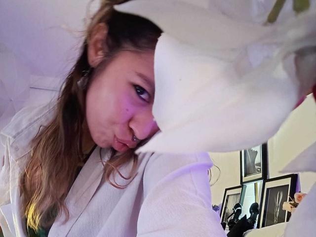 DulceBom with puffy nose, nose ring, and long brown hair, pouting, wearing a white shirt, partially covered by a white cloth, in a slightly blurred selfie.