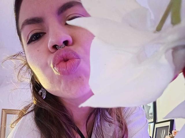 DulceBom with pink nose ring and dark eyeliner, puckering lips near a large white flower, wearing white top, brown hair, and diamond earrings, in a selfie.