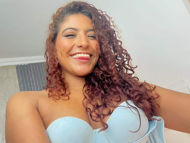 Mia Bianchi mit lockigem, rotem Haar, lächelnd, trägt einen hellblauen BH ohne Träger und zeigt ihre Brüste. Sie hat rosafarbenen Lippenstift und einen süßen, selbstbewussten Ausdruck..