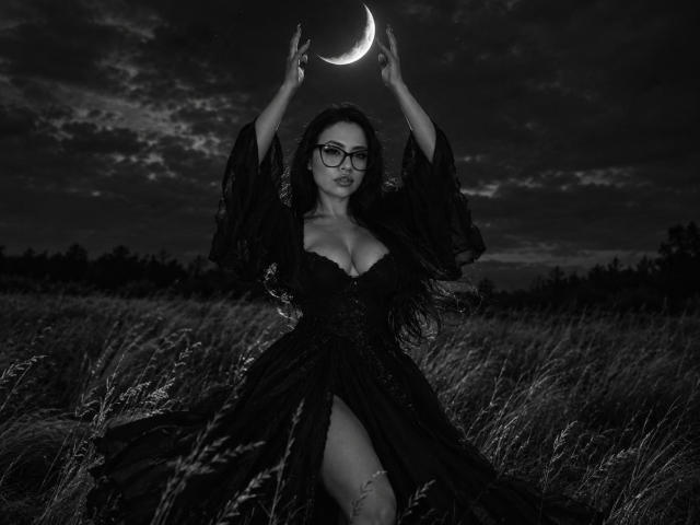 RousseBella with big tits, wearing a black lace dress, raising arms to a crescent moon in a dark, grassy field, wearing glasses, long black hair, nighttime, black and white.