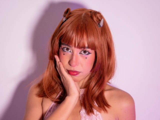 MiluKiss met oranje haar, kattenoren en roze make-up, die haar gezicht aanraakt en haar borsten laat zien, op een foto met roze verlichting, en die er schattig en sexy uitziet..