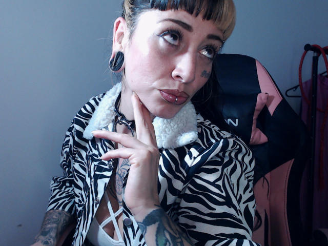 LusstCruella with zebra-print shirt, white fur collar, black ear plugs, tattoos, and pierced lip, looking up with hand on chin, in a pink and black chair.