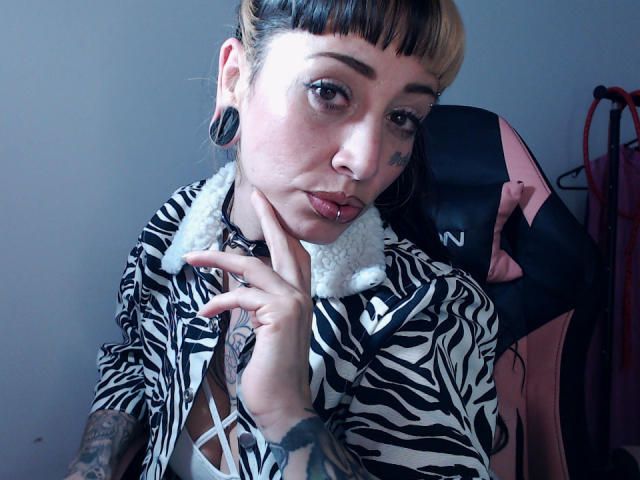 LusstCruella with black and blonde hair, black ear gauges, nose ring, lip ring, tattooed arm, zebra shirt, white fur collar, hand near face, sitting in pink and black chair.