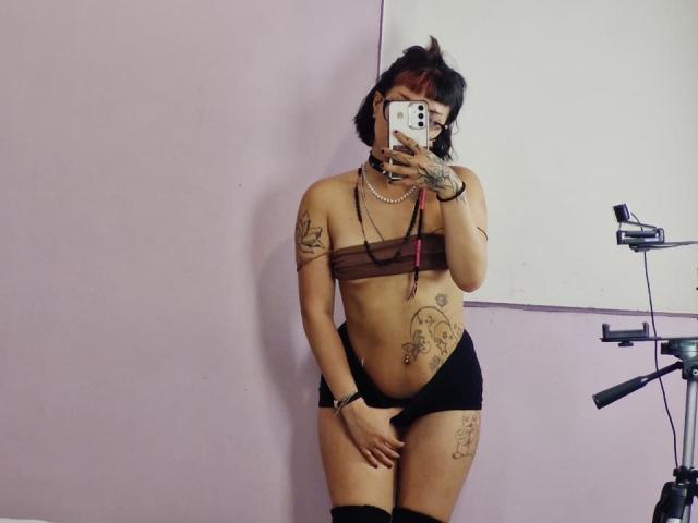 JadeMilleer with dark hair, glasses, and tattoos, wearing a brown strapless top, black shorts, and thigh-high socks, taking a mirror selfie, showing her tits and pussy.