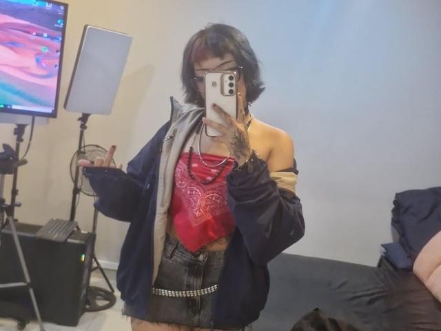JadeMilleer with short black hair, wearing a red bandana top, black shorts, and an off-shoulder jacket, taking a mirror selfie, showing a peace sign, with a TV and light.