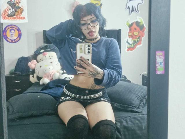 JadeMilleer with blue hair, glasses, and a choker, wearing a blue crop top, black shorts, and thigh-highs, sitting on a bed, sticking out her tongue, showing tattoos.