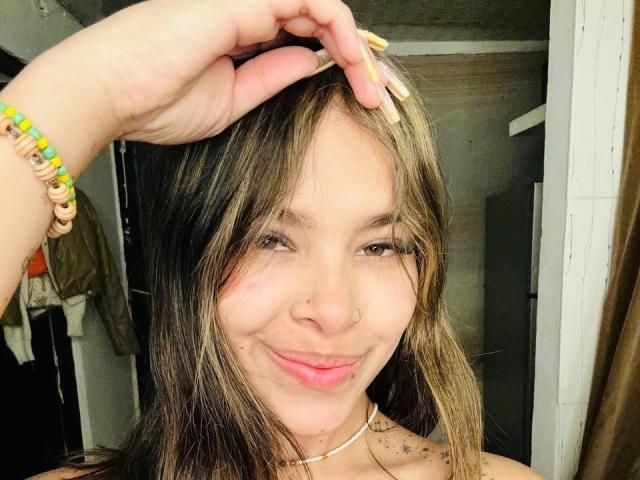 Adhara, con capelli castano scuro, sorridente, con un anello al naso, labbra rosa, tatuaggi a forma di stella, una collana choker, un braccialetto colorato, una mano tra i capelli, pelle chiara, un selfie casuale, illuminazione soffusa, leggero rossore..
