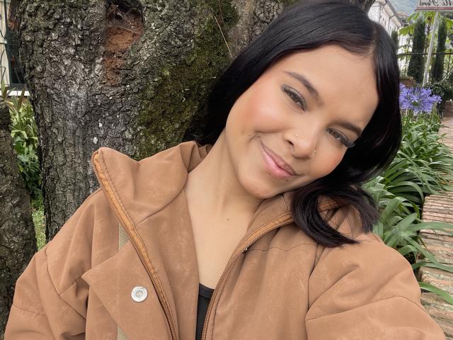 AdharaCopper with black hair, light skin, and a cute smile, wearing a brown suede jacket, has a small nose button and a subtle sheen on her face.