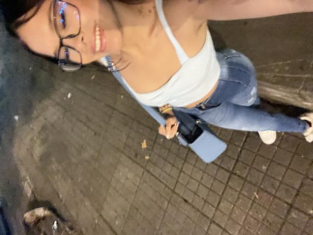 Adhara, mit Brille, lächelnd, trägt ein weißes Tanktop, das ihre kleinen Brüste zeigt, und enge blaue Jeans. Sie steht auf einem Kopfsteinpflasterweg und hält eine blaue Tasche..