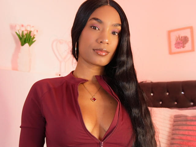 NatLewis with long black hair, wearing a deep red zip-up top, showing her cleavage, wearing a red necklace, and hoop earrings, smiling with pink eyeshadow.